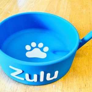 以下が含まれることがあります： 白い肉球のプリントと「Zulu」という名前が側面に印刷された青いシリコン製のペットボウル。ボウルには、簡単に餌を与えられるように注ぎ口が付いています。
