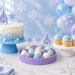 May include: A round cake with blue and white ombre frosting, topped with castle, snowflake, and crown picks. A purple tray holds blue and white cake balls with similar picks. Three cupcakes with blue frosting feature matching decorative toppers.
