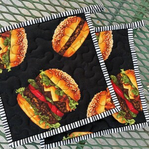 May include: Two square quilted pot holders featuring a black background with a repeating pattern of cheeseburgers. Each pot holder has a black and white striped fabric border and a small fabric loop at the top corner for hanging storage.