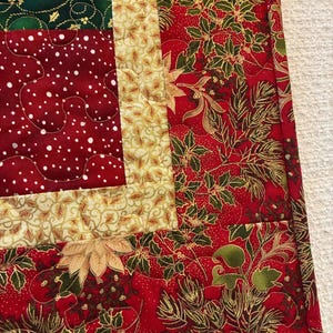 K&ouml;nnte beinhalten: Ein gesteppter Weihnachtstischl&auml;ufer mit Patchwork-Design. Der L&auml;ufer zeigt eine Kombination aus roten, gr&uuml;nen und goldenen Stoffen mit festlichen Mustern, darunter Weihnachtsstern, Stechpalme und Tupfen. Die Kanten sind mit einem passenden Rand sauber verarbeitet.