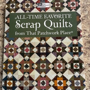 May include: A softcover book titled All-Time Favorite Scrap Quilts from That Patchwork Place. The cover features a repeating geometric quilt pattern in various colors including red, blue, yellow, and brown, with a dark green title block.