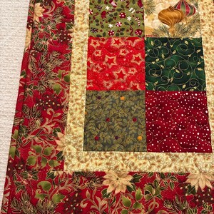 K&ouml;nnte beinhalten: Eine Weihnachtsdecke mit Patchwork-Design. Die Decke zeigt Quadrate mit festlichen Mustern wie Sternen, Ornamenten und Stechpalmen. Die Farbpalette umfasst Rot, Gr&uuml;n und Gold und erzeugt eine warme Urlaubsstimmung. Die R&auml;nder haben ein Blumenmuster.