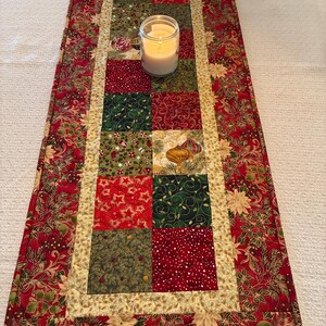 K&ouml;nnte beinhalten: Festlicher Weihnachtstischl&auml;ufer mit Patchwork-Design. Die Mitte zeigt Quadrate aus roten, gr&uuml;nen und goldenen Stoffen mit festlichen Mustern. Eine brennende Kerze in einem Glas steht auf dem L&auml;ufer, der von einem roten Blumendruck umrandet ist.