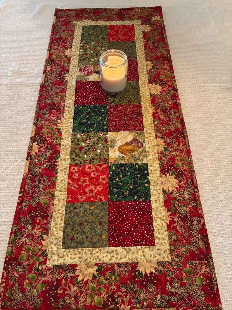 K&ouml;nnte beinhalten: Festlicher Weihnachtstischl&auml;ufer mit Patchwork-Design. Die Mitte zeigt Quadrate aus roten, gr&uuml;nen und goldenen Stoffen mit festlichen Mustern. Eine brennende Kerze in einem Glas steht auf dem L&auml;ufer, der von einem roten Blumendruck umrandet ist.