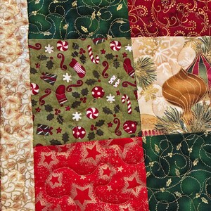 K&ouml;nnte beinhalten: Nahaufnahme eines Patchwork-Quilts mit Weihnachtsmotto. Der Quilt zeigt Stoffquadrate in Rot, Gr&uuml;n und Beige mit Mustern wie Sternen, Zuckerstangen und Ornamenten. Die Steppn&auml;hte sind sichtbar und verleihen Textur.