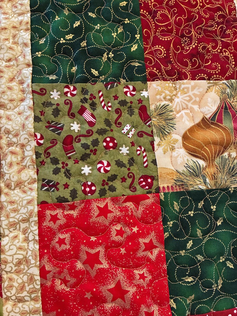 K&ouml;nnte beinhalten: Nahaufnahme eines Patchwork-Quilts mit Weihnachtsmotto. Der Quilt zeigt Stoffquadrate in Rot, Gr&uuml;n und Beige mit Mustern wie Sternen, Zuckerstangen und Ornamenten. Die Steppn&auml;hte sind sichtbar und verleihen Textur.