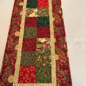 K&ouml;nnte beinhalten: Ein rechteckiger Weihnachtstischl&auml;ufer. Der L&auml;ufer hat ein Patchwork-Design mit roten, gr&uuml;nen und goldenen Stoffen. Die Mitte hat eine Reihe von Quadraten mit festlichen Mustern, die von einem goldenen Rand und einem roten Blumendruck umrahmt sind.
