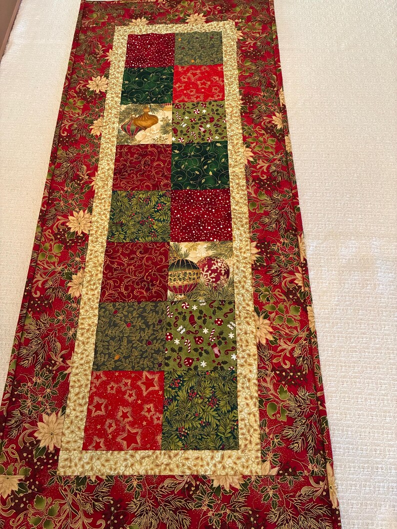 K&ouml;nnte beinhalten: Ein rechteckiger Weihnachtstischl&auml;ufer. Der L&auml;ufer hat ein Patchwork-Design mit roten, gr&uuml;nen und goldenen Stoffen. Die Mitte hat eine Reihe von Quadraten mit festlichen Mustern, die von einem goldenen Rand und einem roten Blumendruck umrahmt sind.