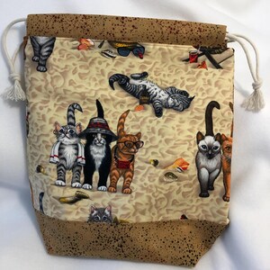 May include: A brown drawstring bag with a beige fabric lining featuring a repeating pattern of cats wearing sunglasses, hats, and other accessories on a beach scene.