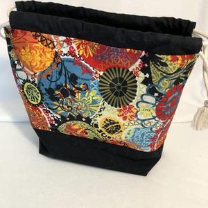 May include: A fabric drawstring bag with a colorful floral pattern in red, orange, yellow, and blue. The bag has a black base and top trim, with a white drawstring closure. The bag is designed to stand upright.