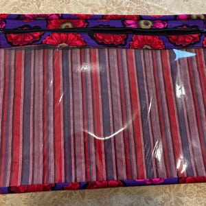 May include: A rectangular storage bag with a clear front and a floral and striped design. The top edge features a purple fabric with red flowers and a black zipper. The main body has vertical red, purple, and white stripes.