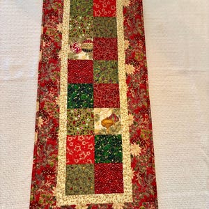 May include: A rectangular Christmas table runner with a patchwork design. The center features squares of red, green, and gold fabrics with festive patterns. The border is red with a floral design, and the inner border is gold.