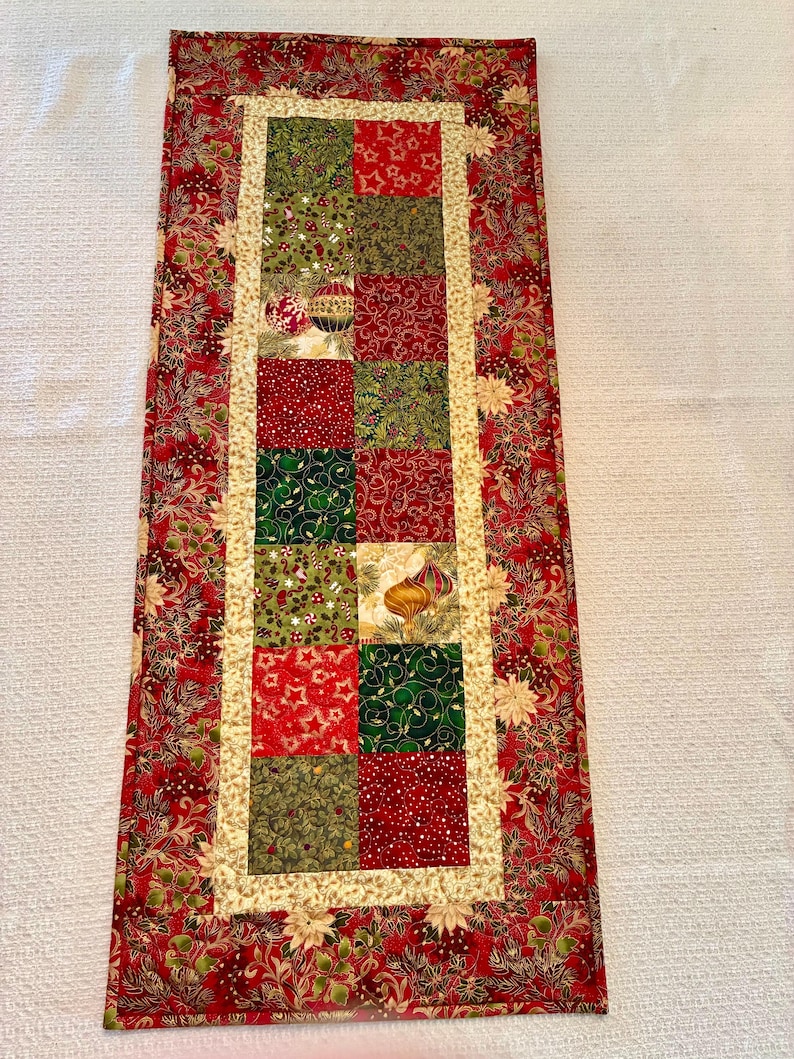 K&ouml;nnte beinhalten: Ein rechteckiger Weihnachtstischl&auml;ufer mit Patchwork-Design. Die Mitte zeigt Quadrate aus roten, gr&uuml;nen und goldenen Stoffen mit festlichen Mustern. Der Rand ist rot mit einem floralen Design, der innere Rand ist goldfarben.