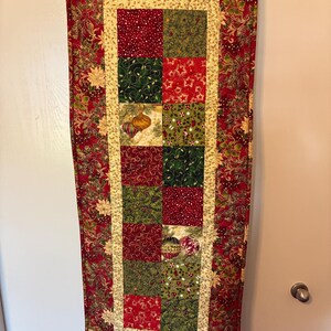 K&ouml;nnte beinhalten: Ein rechteckiger Weihnachtstischl&auml;ufer. Der L&auml;ufer hat ein Patchwork-Design mit roten, gr&uuml;nen und goldenen Quadraten. Der Rand ist tiefrot mit einem floralen Muster. Die mittleren Quadrate haben Weihnachtsschmuck und Sternmuster.