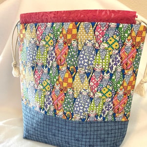May include: A drawstring bag featuring a vibrant, multi-colored cat pattern with geometric shapes in shades of blue, green, yellow, and pink. The base is a blue fabric with a dark grid pattern, and the top edge is a solid, textured magenta fabric.