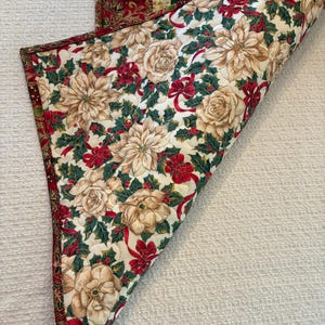 K&ouml;nnte beinhalten: Ein gesteppter Tischl&auml;ufer zum Thema Weihnachten mit floralem Muster. Der L&auml;ufer zeigt cremefarbene Blumen, rote Schleifen und gr&uuml;ne Stechpalmenbl&auml;tter auf wei&szlig;em Hintergrund. Die R&auml;nder sind mit einem rot-goldenen Stoff verziert.