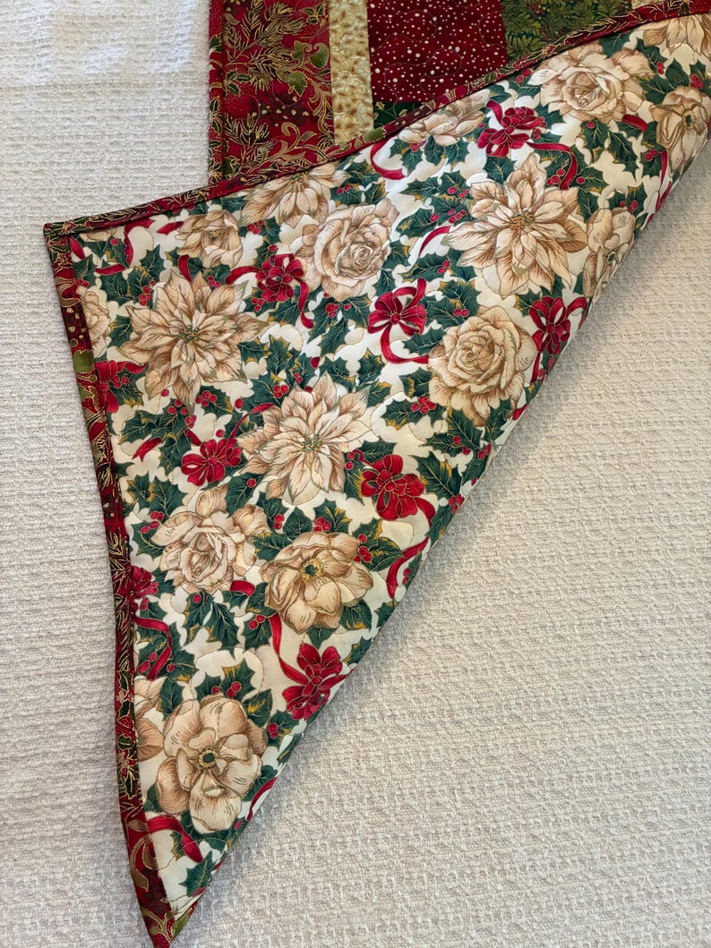 K&ouml;nnte beinhalten: Ein gesteppter Tischl&auml;ufer zum Thema Weihnachten mit floralem Muster. Der L&auml;ufer zeigt cremefarbene Blumen, rote Schleifen und gr&uuml;ne Stechpalmenbl&auml;tter auf wei&szlig;em Hintergrund. Die R&auml;nder sind mit einem rot-goldenen Stoff verziert.