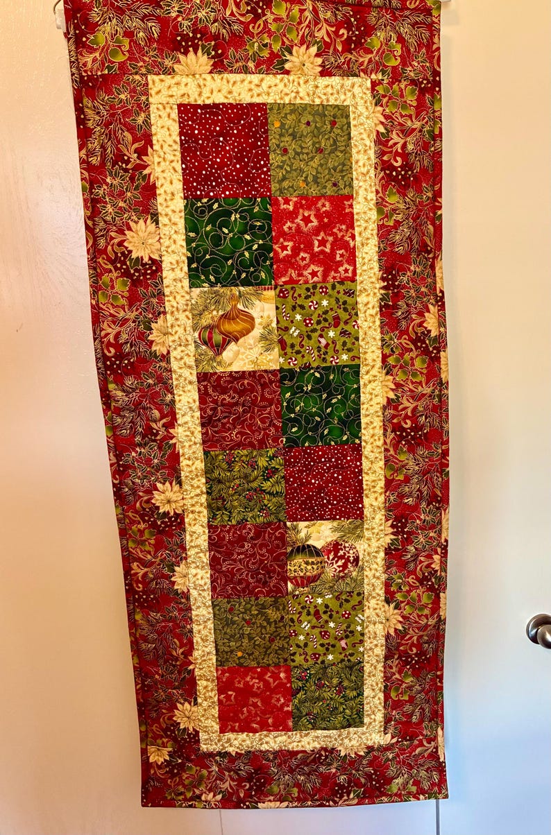 K&ouml;nnte beinhalten: Ein weihnachtliches Tischl&auml;ufer mit Patchwork-Design. Der L&auml;ufer hat einen roten Hintergrund mit floralen Mustern und ein zentrales Feld aus roten, gr&uuml;nen und goldenen Quadraten, die mit Weihnachtsschmuck und festlichen Motiven verziert sind.
