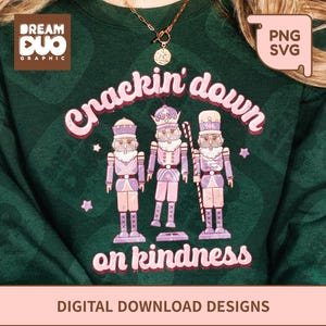 May include: Dark green sweatshirt featuring a graphic of three nutcrackers in pink and purple, with the words "Crackin' down on kindness". The sweatshirt also has the Dream Duo Graphic logo and a PNG SVG tag.