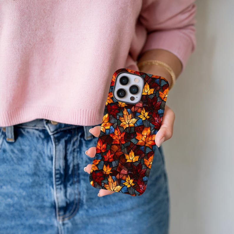 May include: A smartphone case with a stained-glass effect, displaying a pattern of autumn leaves in red, orange, yellow, and blue. The case is held in a hand, highlighting the vibrant autumnal design. The case is designed for a smartphone.