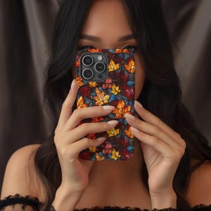 May include: A smartphone case with a vibrant autumn leaf pattern. The case showcases a mosaic of red, yellow, and orange leaves on a dark backdrop. The phone case is held in front of a person's face.