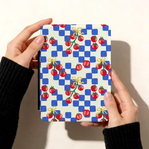 May include: A patterned case featuring a blue and white checkered background with illustrations of red tomatoes on vines tied with yellow bows. The case is held by two hands.