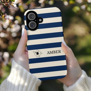 May include: A smartphone case with a blue and white striped pattern. The case features the name "AMBER" in black text, along with a small floral graphic. The case has a camera cutout at the top.