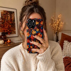 May include: A smartphone case with a vibrant autumn leaf pattern in red, orange, yellow, and blue. The case has a glossy finish and a black edge. The phone case is held up in front of a person's face.