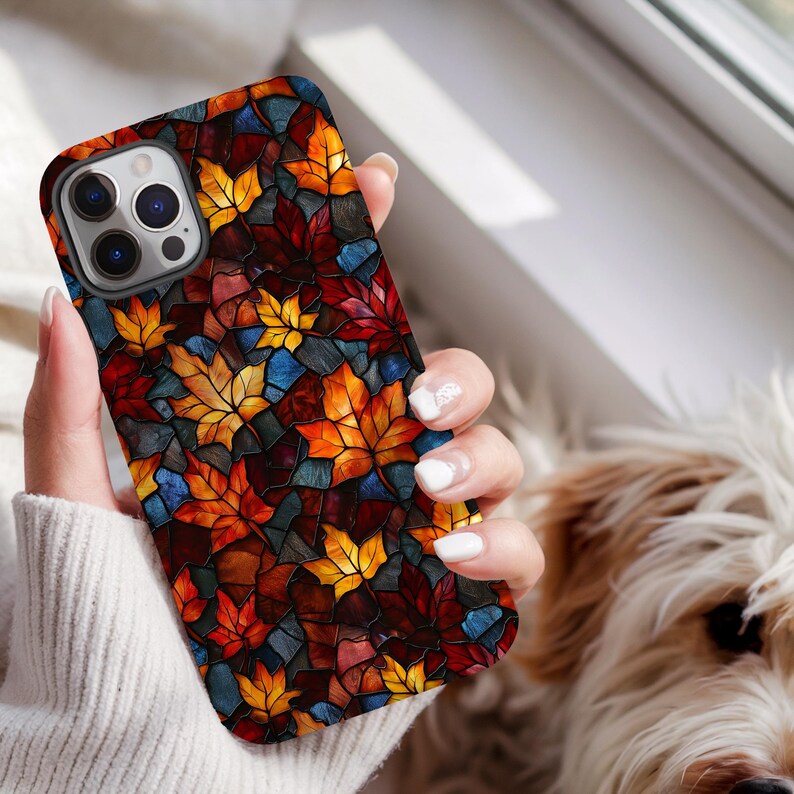 May include: A smartphone case with a stained-glass effect, depicting autumn leaves in red, orange, yellow, and blue. The case has a shiny finish and a dark border. The phone case is held by a person wearing a white jumper.