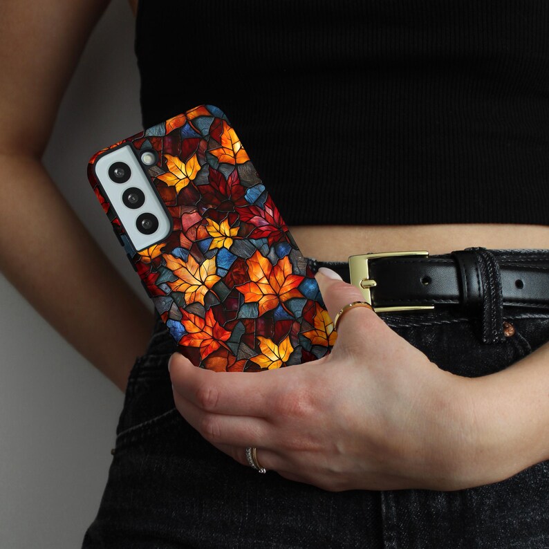 May include: A mobile phone case with a stained-glass effect, displaying a pattern of autumn leaves in orange, red, yellow, and blue. The case is held in a hand, highlighting the decorative design. The phone accessory is a protective cover.
