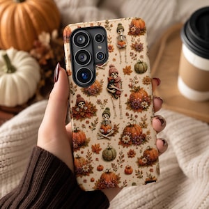 May include: A beige smartphone case featuring a repeating pattern of skeletons wearing knit hats, orange pumpkins, autumn leaves, and small flowers. The design includes skeletons holding pumpkins or black cats, set against a light cream-colored background.