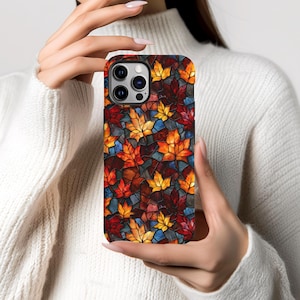 May include: A phone case with a stained glass effect, depicting autumn leaves in red, orange, yellow, and blue. The case has a glossy finish and a dark border. The phone case is held in a person's hands.