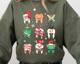 以下が含まれることがあります： クリスマスをテーマにした緑色のスウェットシャツ。漫画の歯が、お祝いの帽子やアクセサリーを身に着けています。歯は、キャンディーケーン、雪片、クリスマスツリーで飾られています。