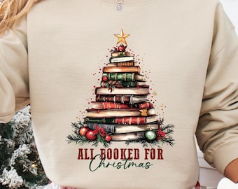 以下が含まれることがあります： 本で作られたクリスマスツリーのデザインが施されたベージュのスウェットシャツ。星とオーナメントで飾られています。「ALL BOOKED FOR Christmas」の文字が下にプリントされています。赤と白のチェック柄のパジャマパンツを履き、サンタクロースのイラストが描かれたマグカップを持っています。