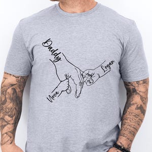 May include: Gray t-shirt with a black line drawing of two hands holding each other. The text "Daddy" is above the larger hand, "Nova" is below the smaller hand, and "Logan" is above the smaller hand.
