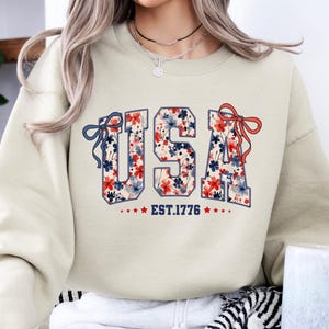 May include: A beige sweatshirt with a floral print of the word "USA" and the text "EST. 1776" below it.