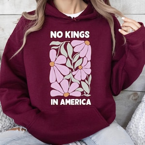 May include: Burgundy hoodie with a floral design and the text "NO KINGS IN AMERICA" in white. The graphic features pink flowers and green leaves. The hoodie has a front pocket and a hood.