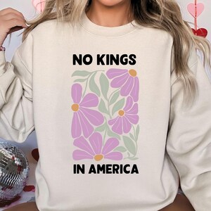 May include: A cream-coloured sweatshirt featuring the text "NO KINGS IN AMERICA" above a floral design of purple flowers and green leaves. The sweatshirt is a casual, comfortable style.
