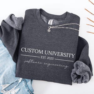 May include: A dark gray sweatshirt with the text "CUSTOM UNIVERSITY EST. 2023 software engineering" in white. A silver necklace with a pendant is draped across the front. The sweatshirt is paired with light blue jeans.