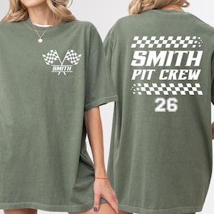 May include: Olive green t-shirt with white graphics. The front features two checkered flags and the name "SMITH" below. The back displays "SMITH PIT CREW" with a checkered pattern and the number "26".