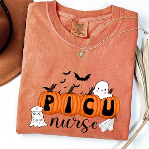 May include: Orange Comfort Colors t-shirt with a Halloween-themed design. The design includes pumpkins, bats, a ghost, and the words "PICU nurse". A gold necklace is draped across the shirt.