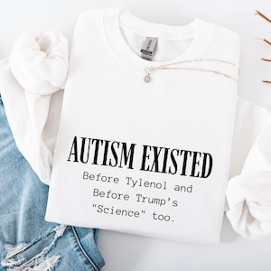 May include: White sweatshirt with the text "AUTISM EXISTED" in bold black letters. Below the text are the words "Before Tylenol and Before Trump's 'Science' too." A silver necklace with a small pendant rests on the sweatshirt.