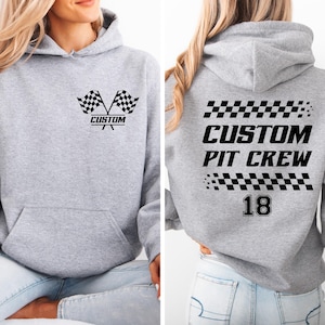 Peut inclure: Un sweat à capuche gris clair avec une poche avant et une capuche. Le devant présente des drapeaux à damier croisés et le mot "CUSTOM". Le dos affiche "CUSTOM PIT CREW" et le numéro "18", le tout en noir. Design sportif et décontracté.