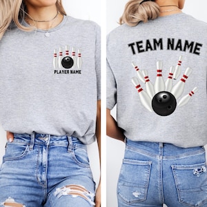 May include: A light gray t-shirt featuring a bowling design. The front displays a bowling ball and pins with "PLAYER NAME" text. The back shows a similar design with "TEAM NAME" text. The shirt is made of a soft material.
