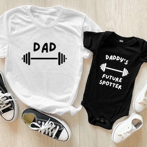 Peut inclure: T-shirt blanc avec le texte "DAD" et un graphique de barre d'haltères. Combinaison noire avec le texte "Daddy's Future Spotter" et un graphique de barre d'haltères.