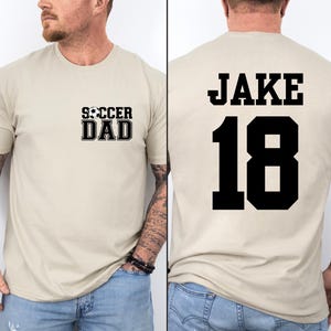 Może przedstawiać: Beżowa koszulka z napisem "Soccer Dad" i grafiką piłki nożnej z przodu. Z tyłu koszulka ma napis "Jake 18" w kolorze czarnym.