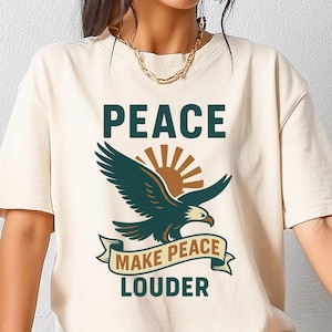 May include: Cream-colored t-shirt featuring the word "PEACE" above a teal eagle graphic with a sunburst and the text "MAKE PEACE LOUDER" on a banner. The shirt has short sleeves and a relaxed fit.