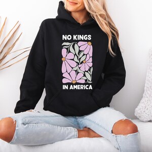 May include: Black hooded sweatshirt with the text "NO KINGS IN AMERICA" in white. The sweatshirt features a floral design in pink, green, and gold. It has a front pocket and a hood.