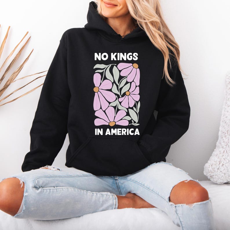 May include: Black hooded sweatshirt with the text "NO KINGS IN AMERICA" in white. The sweatshirt features a floral design in pink, green, and gold. It has a front pocket and a hood.