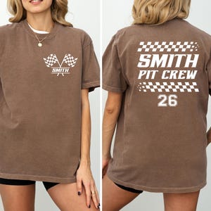 以下が含まれることがあります： レーシングテーマの茶色のTシャツ。前面には、交差したチェッカーフラッグと「SMITH」の文字が描かれています。背面には「SMITH PIT CREW」と数字の「26」、チェッカーフラッグのデザインが表示されています。